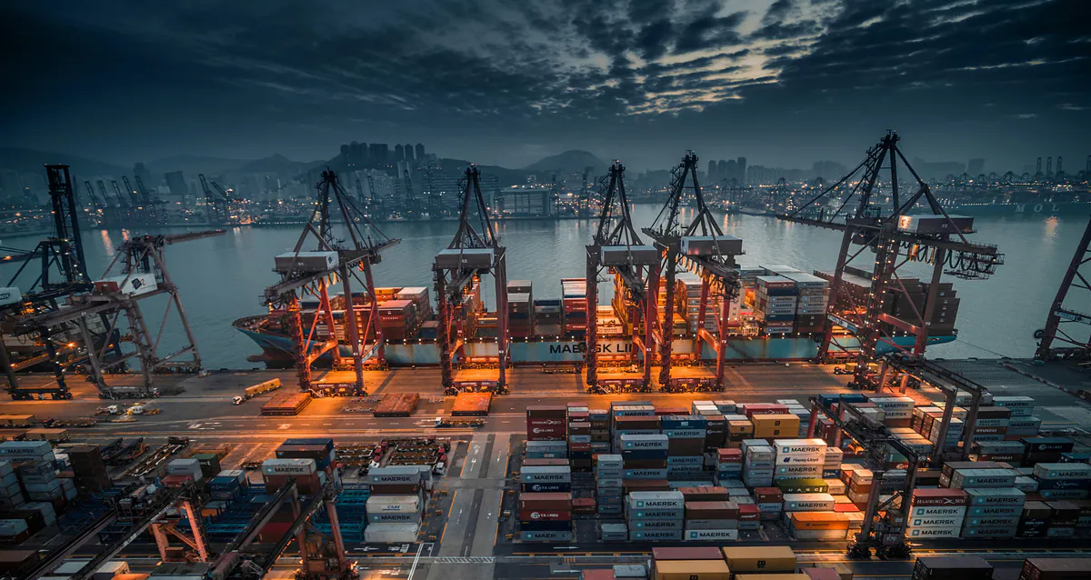 Container cranes loading a cargo ship at an illuminated industrial port in Hong Kong.