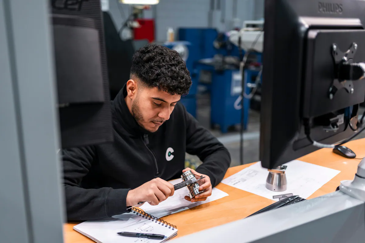 Technician checking a machined metal component with a digital caliper at an engineering workstation.